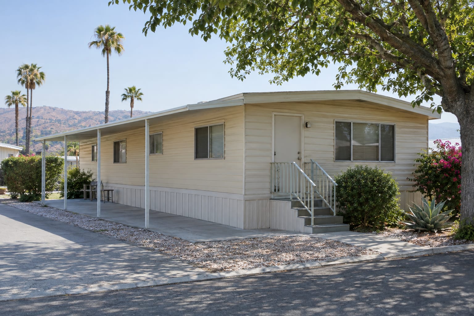An older single-wide mobile home with a nearly flat roof, ribbed metal siding and a long metal awning on a tree-shaded space in an established California community