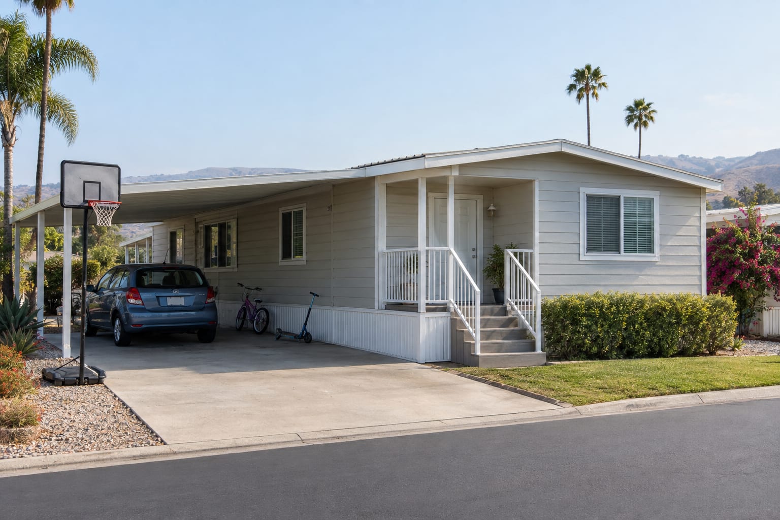 A child's bicycle and a basketball hoop beside the carport of a manufactured home on a community street.