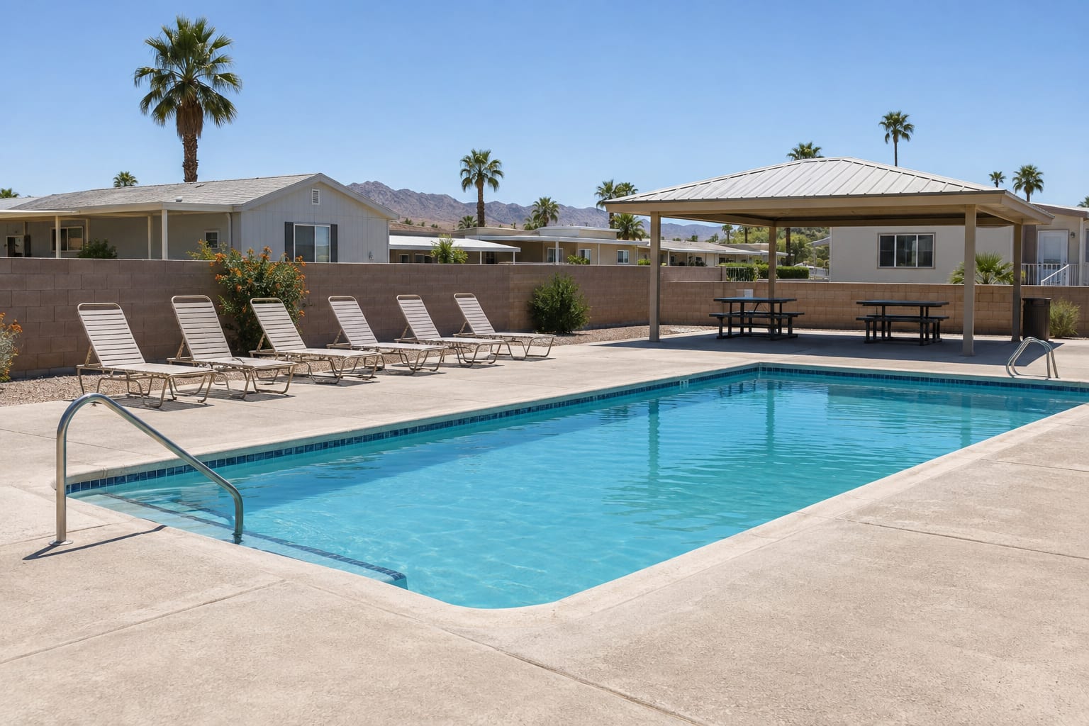 An empty community swimming pool with lounge chairs and a shade ramada, with manufactured homes visible beyond a low wall.