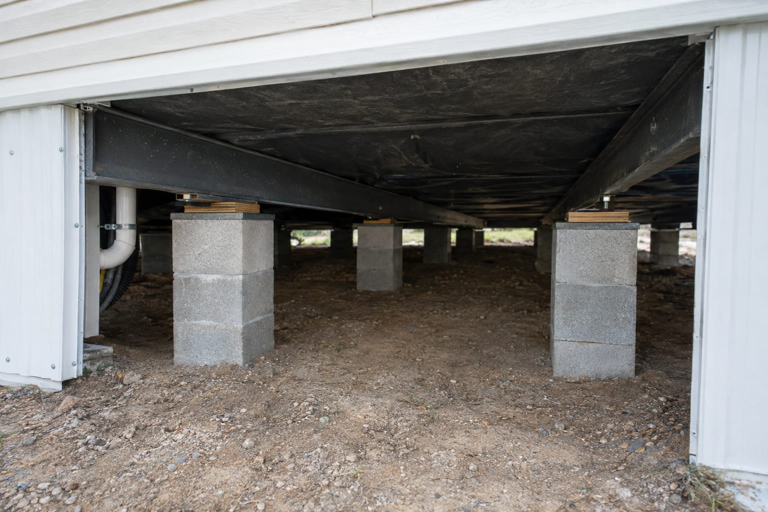 The underside of a manufactured home seen through an open skirting panel: steel frame beams resting on a row of concrete-block piers.
