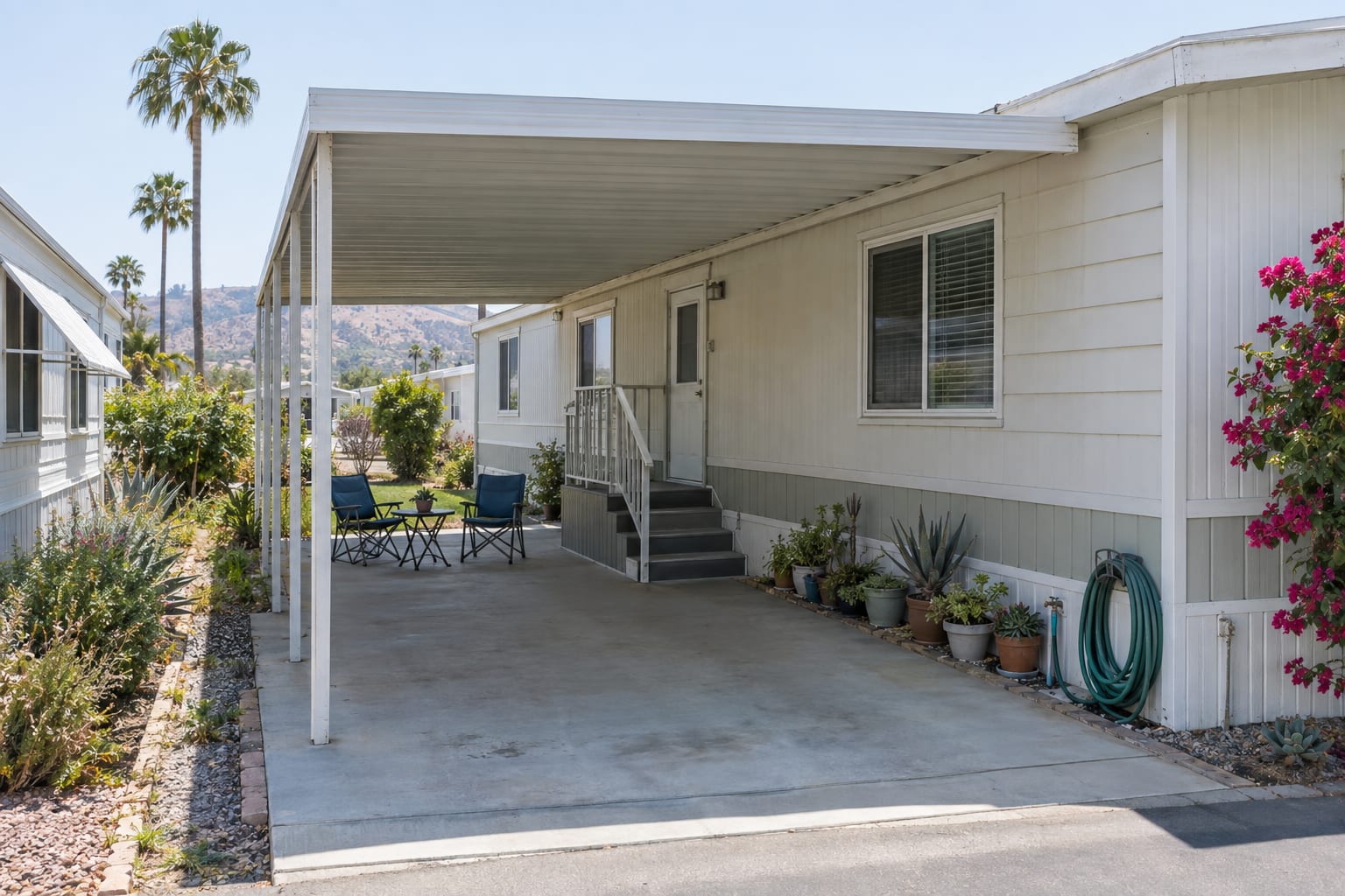 A carport used as a patio beside a single-wide home, with two chairs, potted plants and the neighboring home close by.