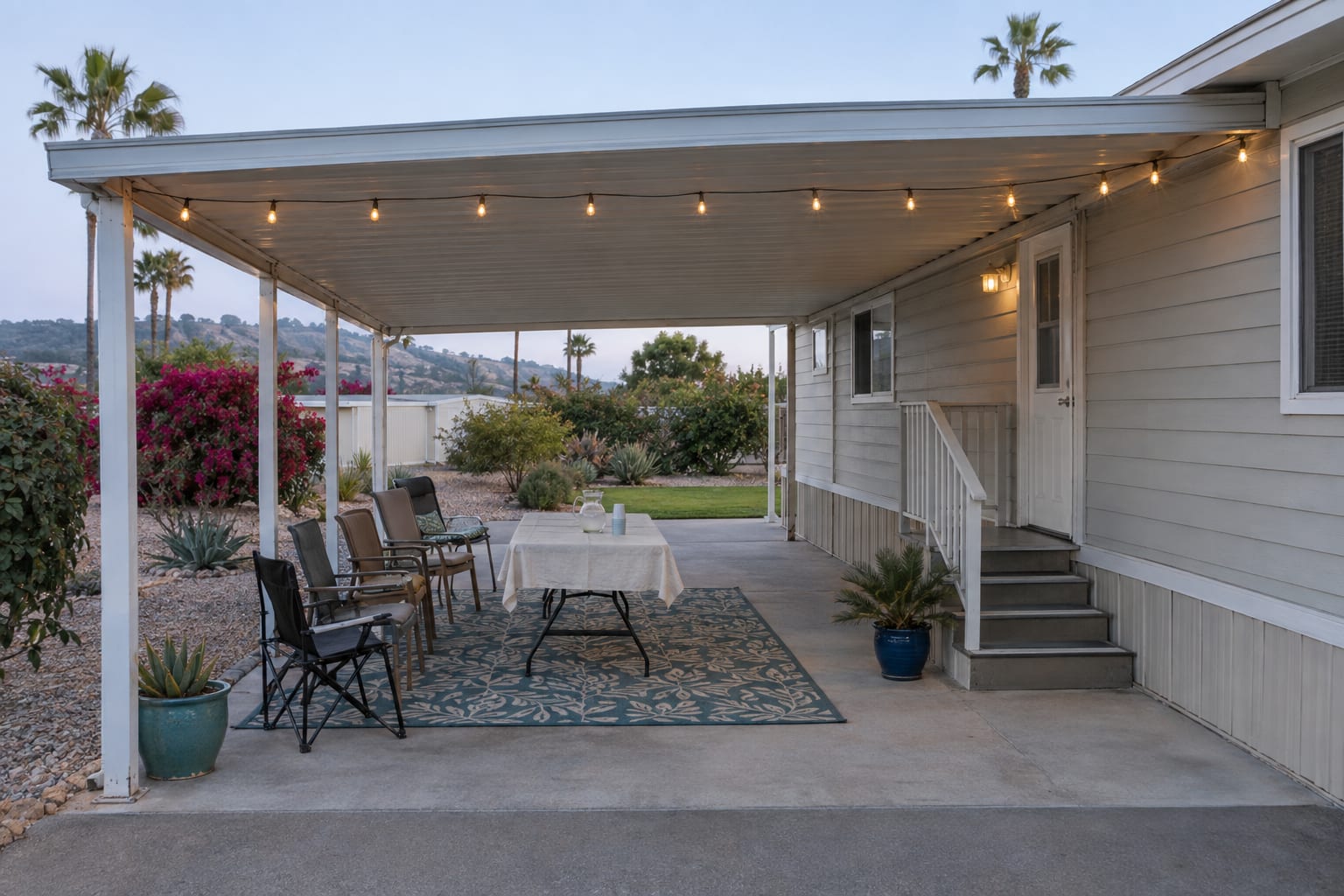 A carport with an outdoor rug, a folding table, chairs and string lights along the beam, beside the side door of a manufactured home.