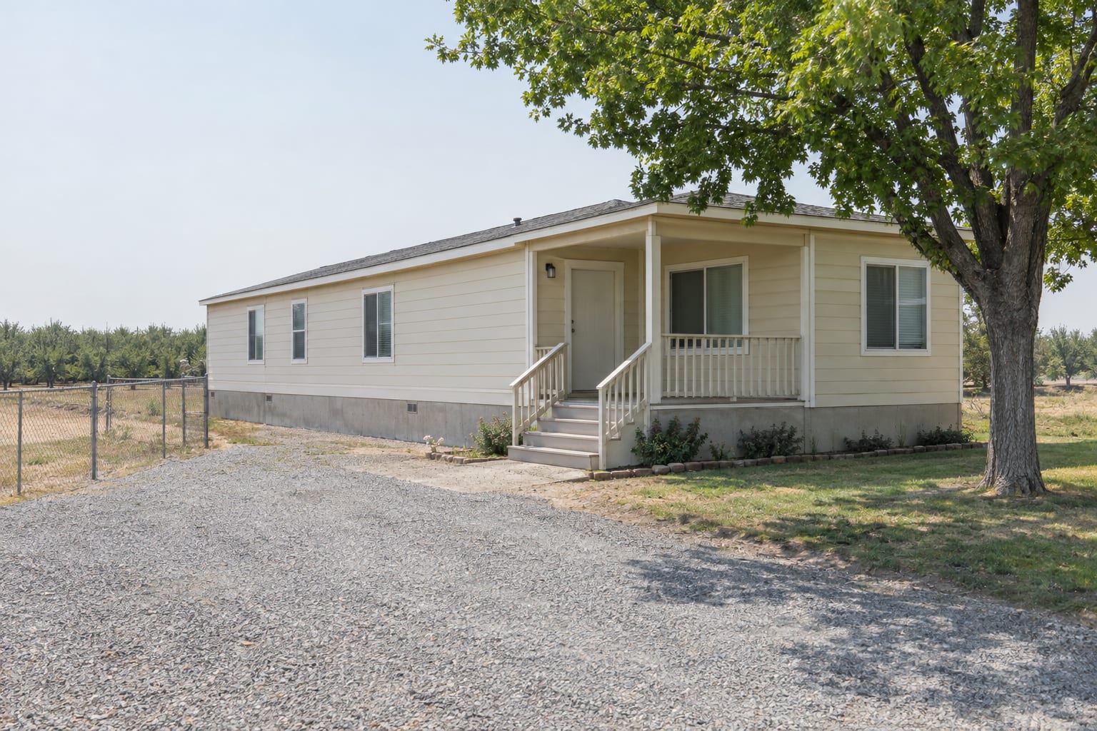 An older double-wide manufactured home on a low foundation on its own flat Central California lot, with a shade tree beside it and orchard rows in the distance