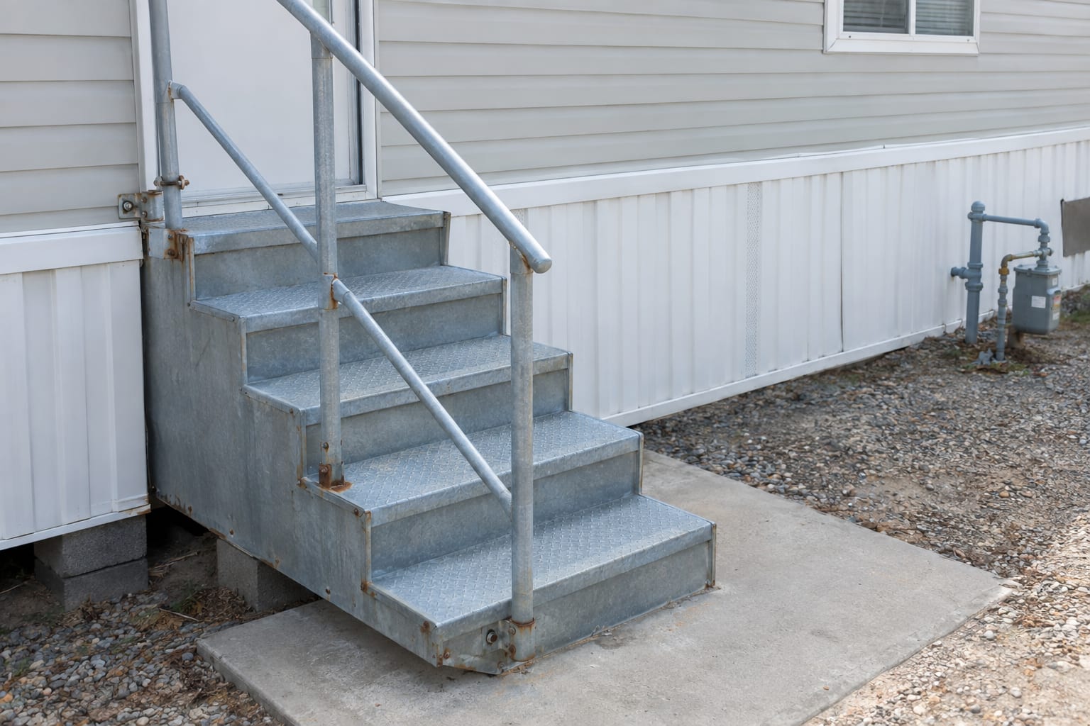A close view of galvanized steel steps and vinyl skirting at the base of a manufactured home in a coastal community, with light surface corrosion on the fasteners