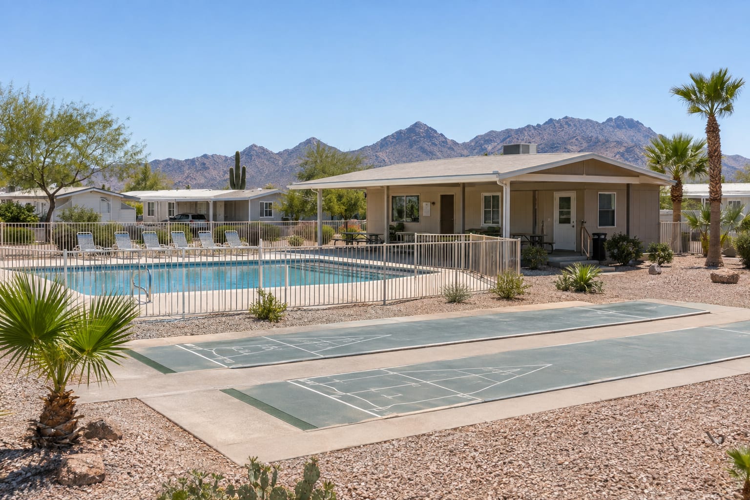 A single-storey community clubhouse with a shaded ramada, a fenced pool and shuffleboard courts, with manufactured homes behind.