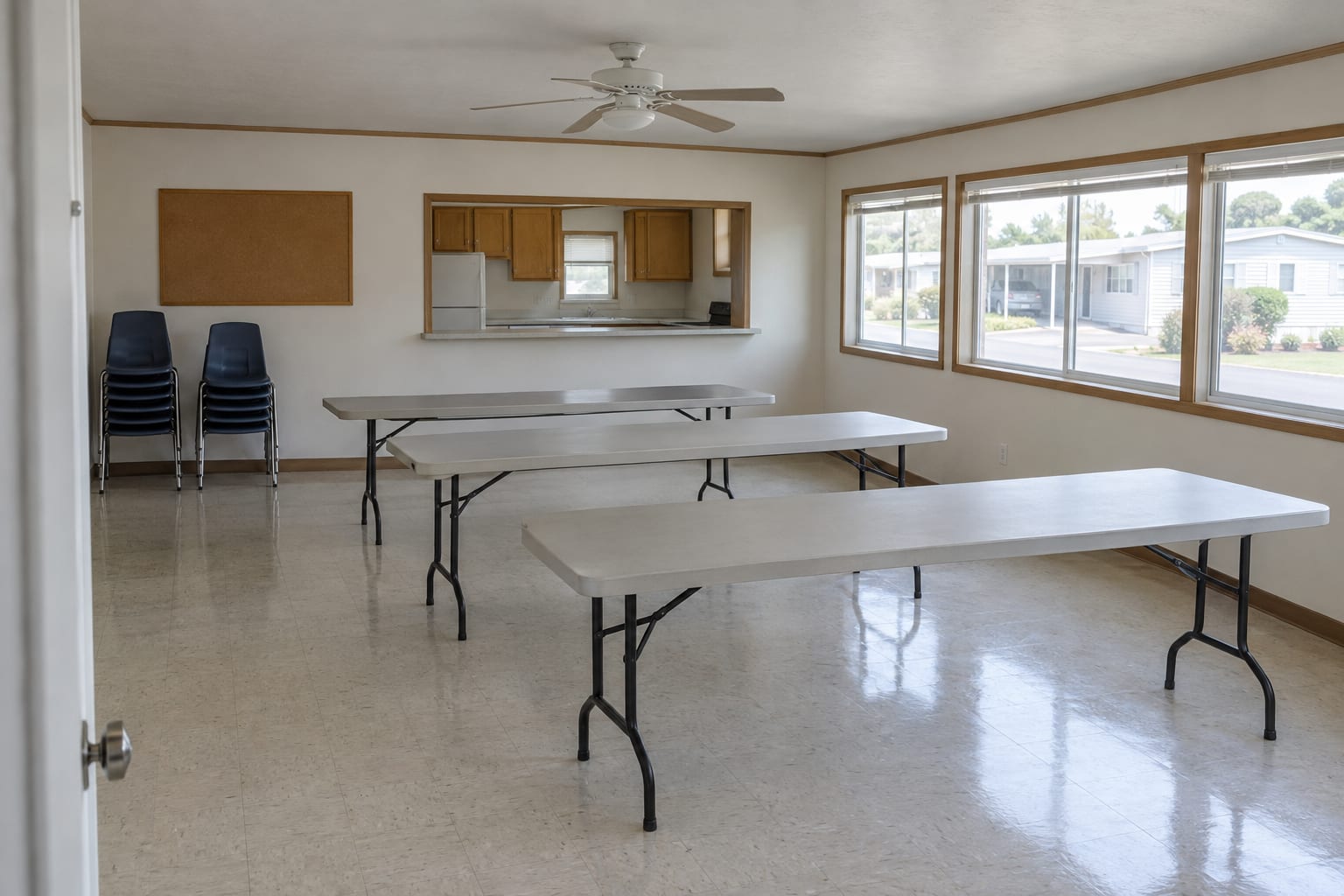 An empty community clubhouse room with folding tables, stacked chairs and a kitchen pass-through, lit by daylight from the windows.