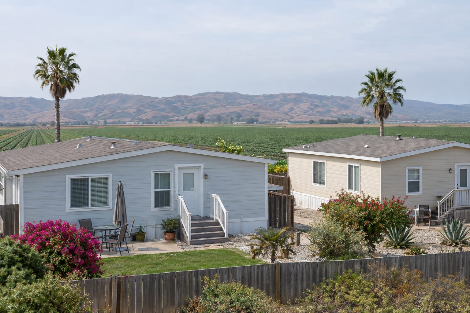 Two double-wide manufactured homes along the edge of a community, with flat fields of row crops and low coastal hills beyond the fence.