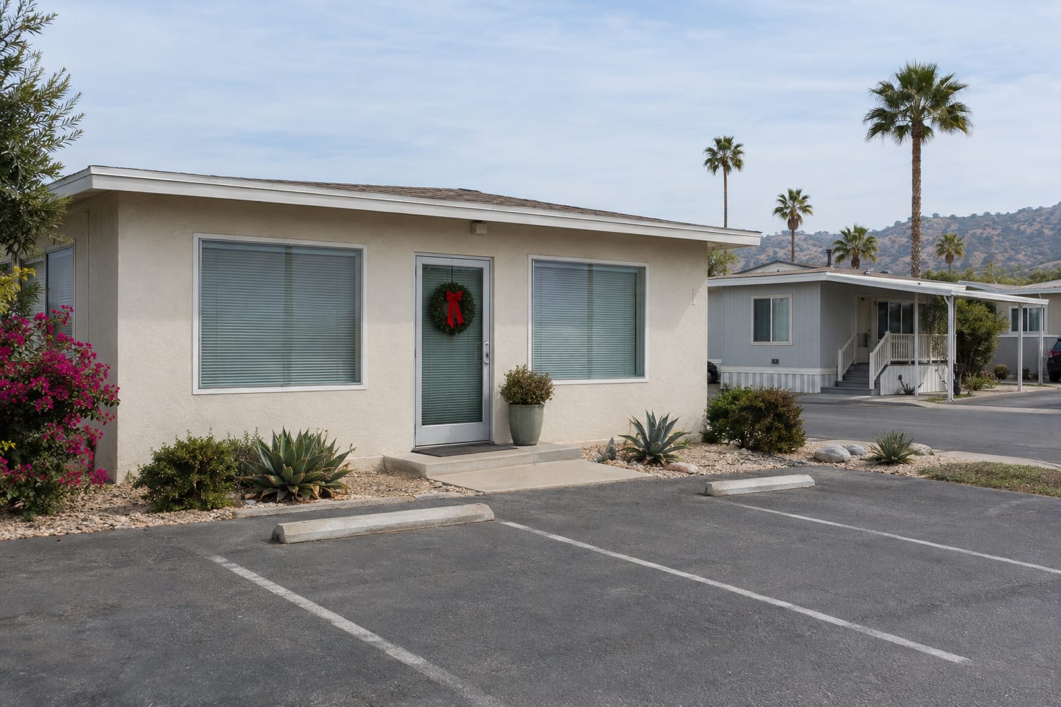 A small community office with a wreath on its closed door, drawn blinds and an empty parking area, with manufactured homes behind it