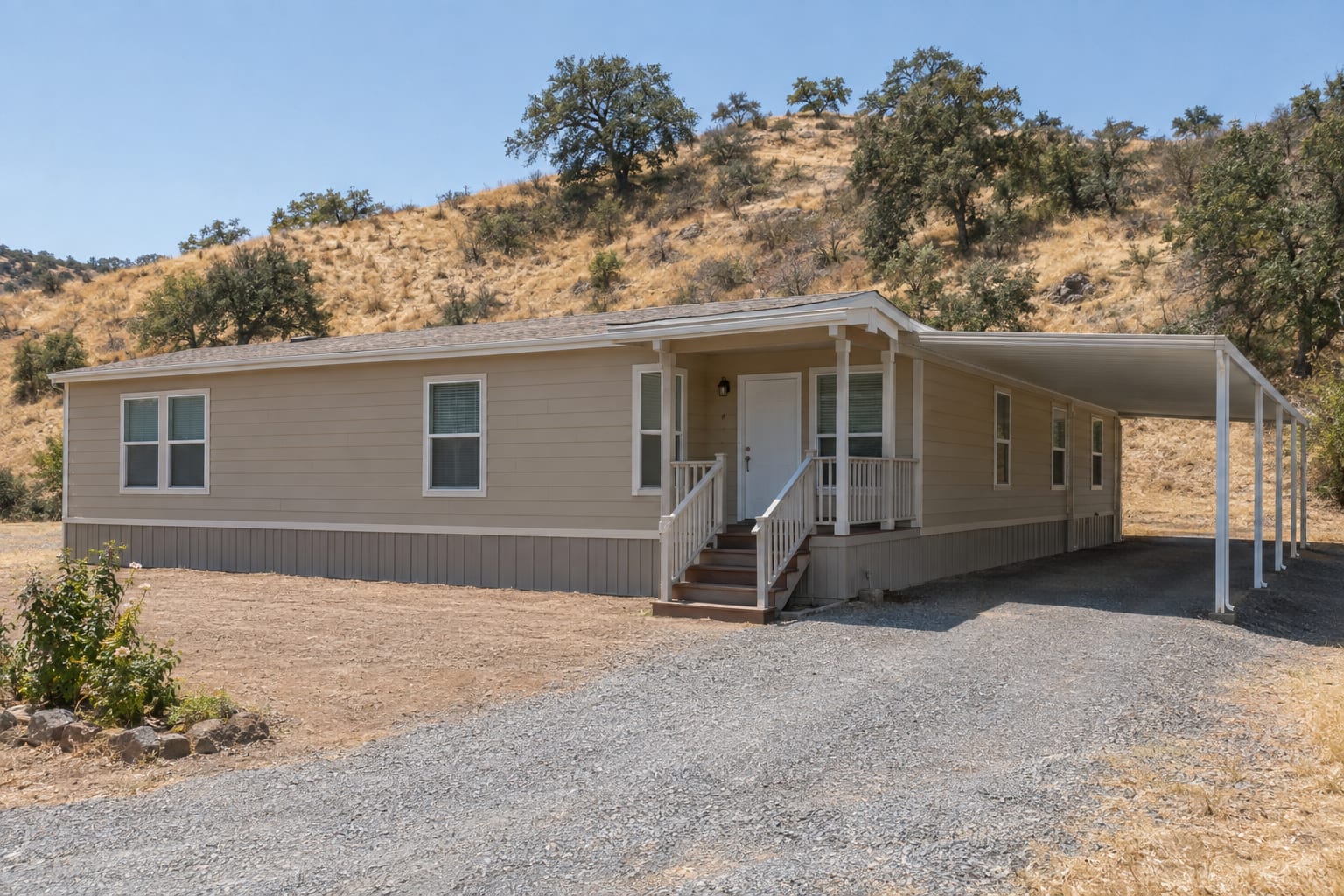 A manufactured home on a lot below a dry brush-covered hillside, with bare cleared ground around it.