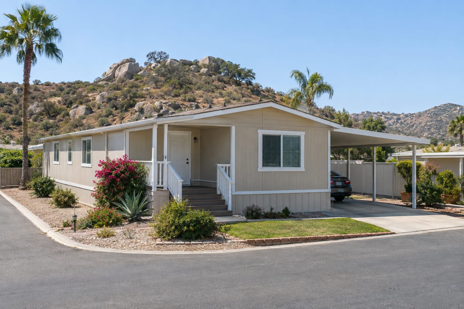A double-wide manufactured home on a corner space at the edge of an inland community, with a boulder-strewn chaparral hillside rising behind it