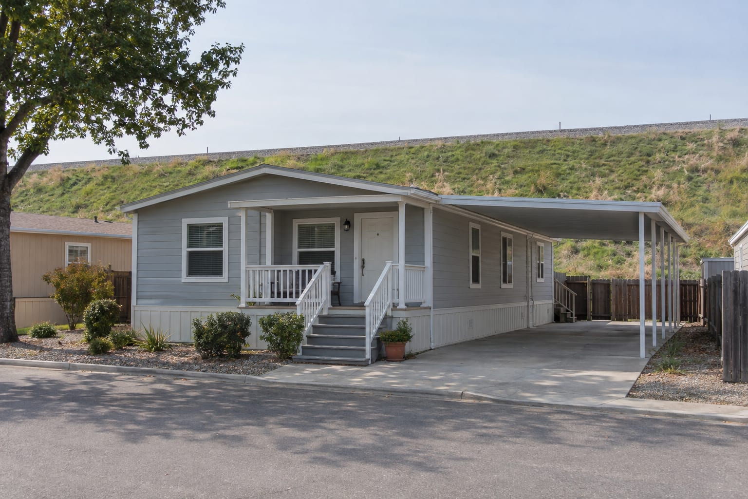 A manufactured home with a carport and a covered porch on a community street, with a high grassy levee embankment rising behind the back fences.