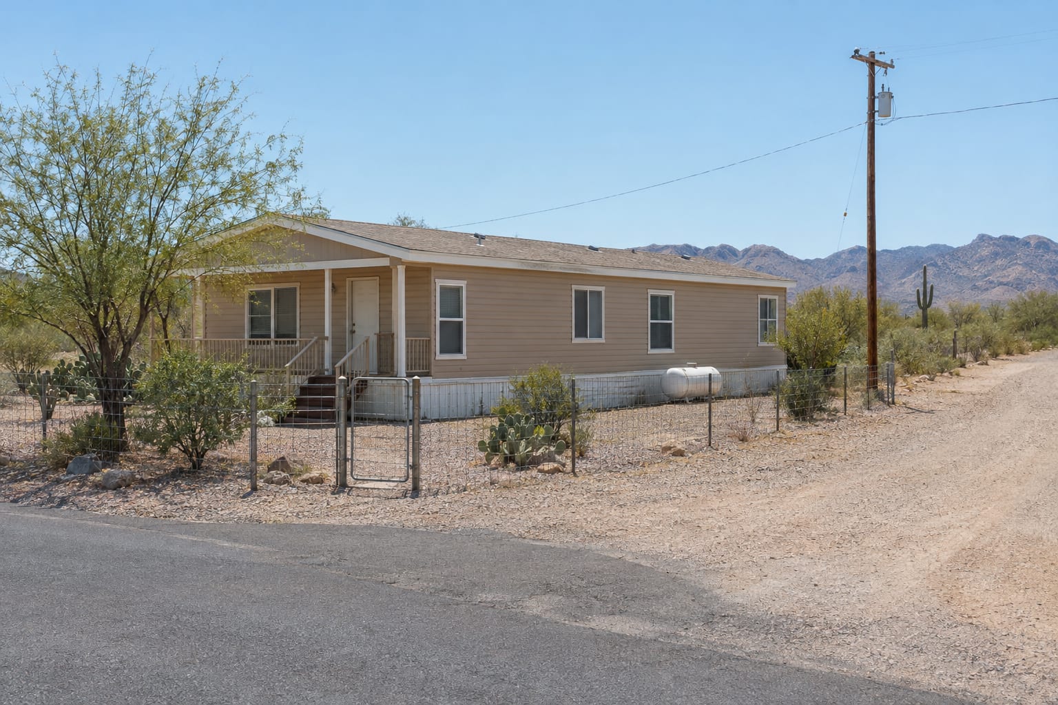 A tan double-wide manufactured home with a propane tank on a fenced lot where a paved street turns into a dirt road at the edge of a small desert town
