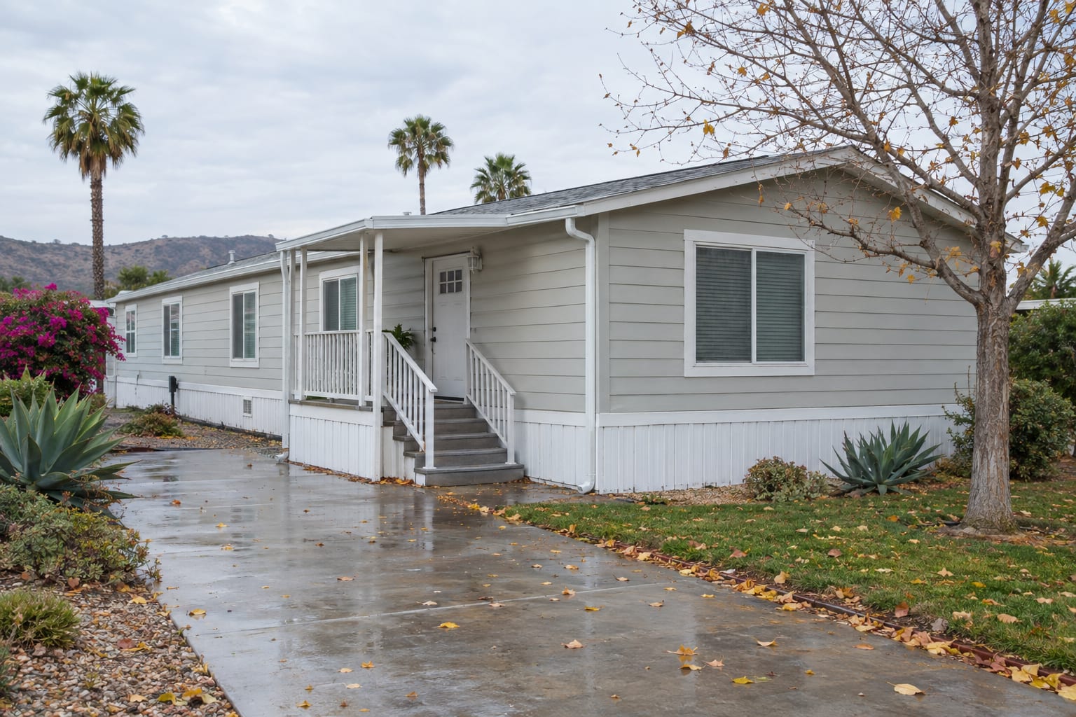 A double-wide manufactured home on an overcast autumn day, with fallen leaves on the wet driveway and along the gutter