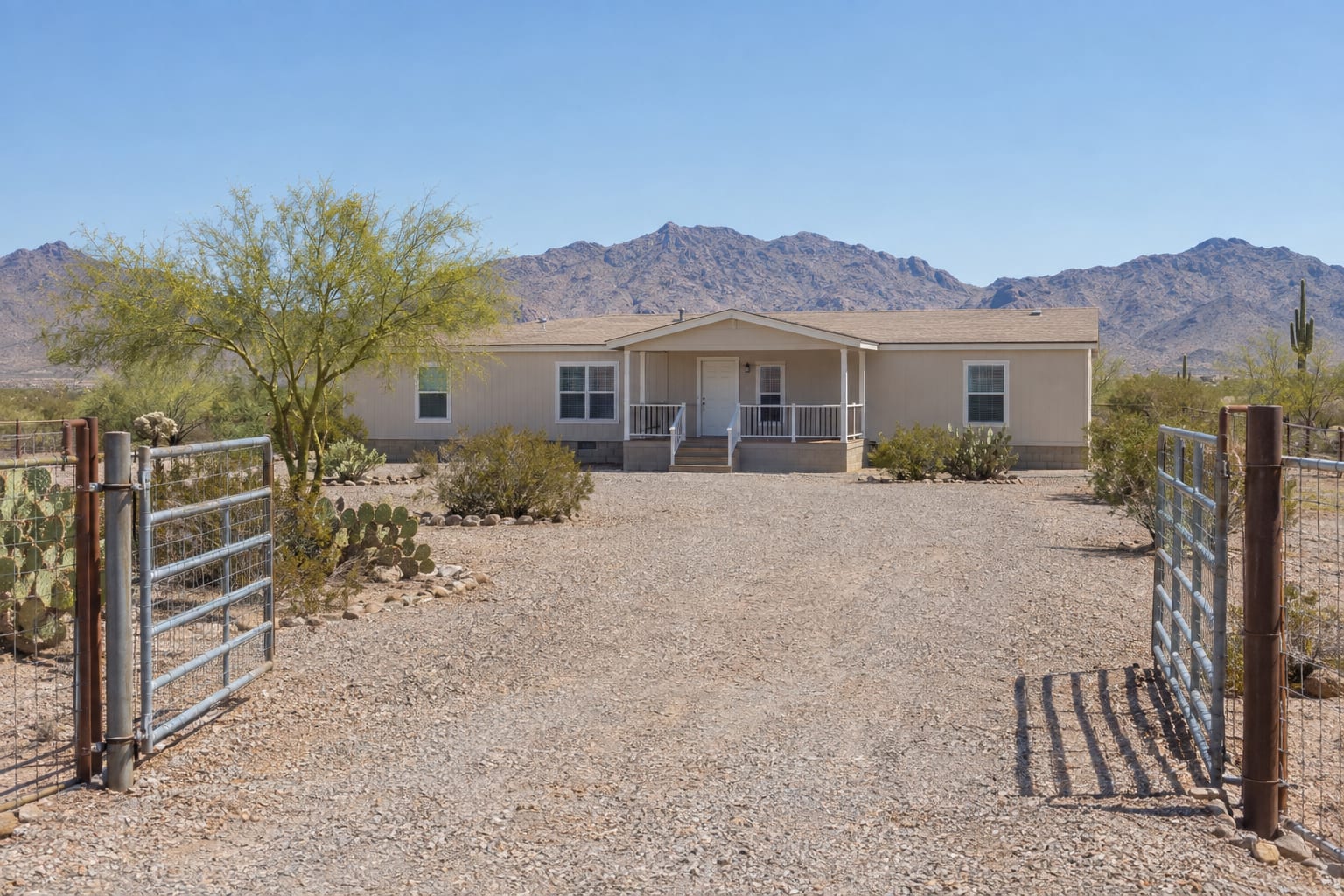 A manufactured home on a fenced desert lot in Arizona, seen from an open gate at the end of a gravel driveway.