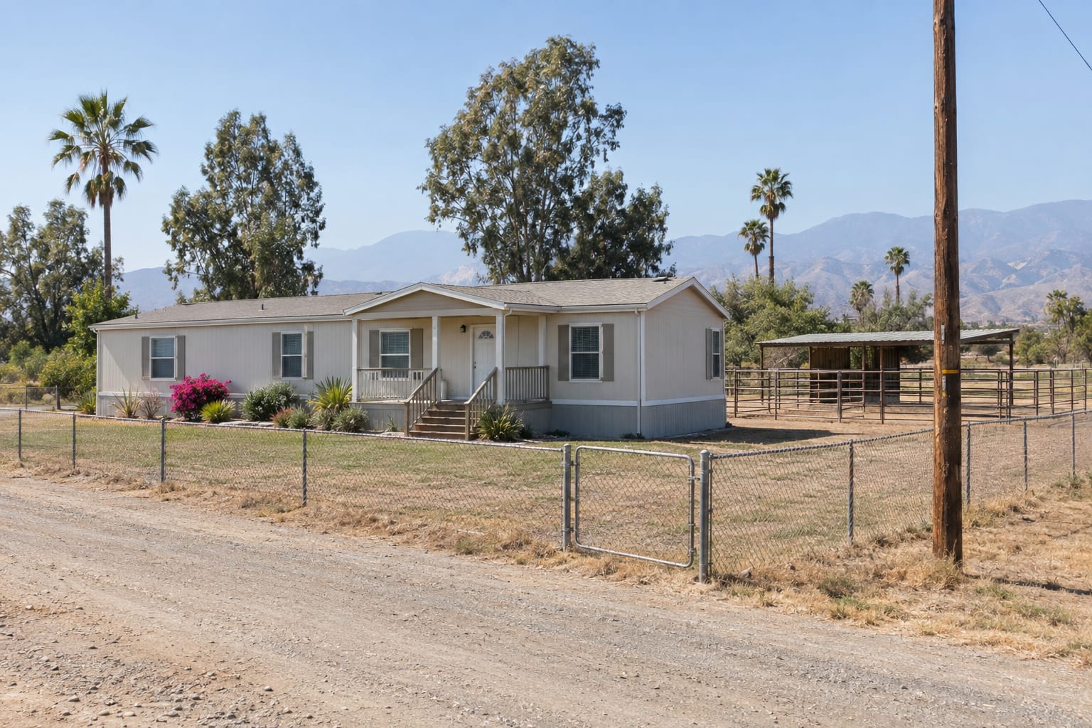 A manufactured home on a large flat lot with a pipe-rail corral beside it and mountains in the distance