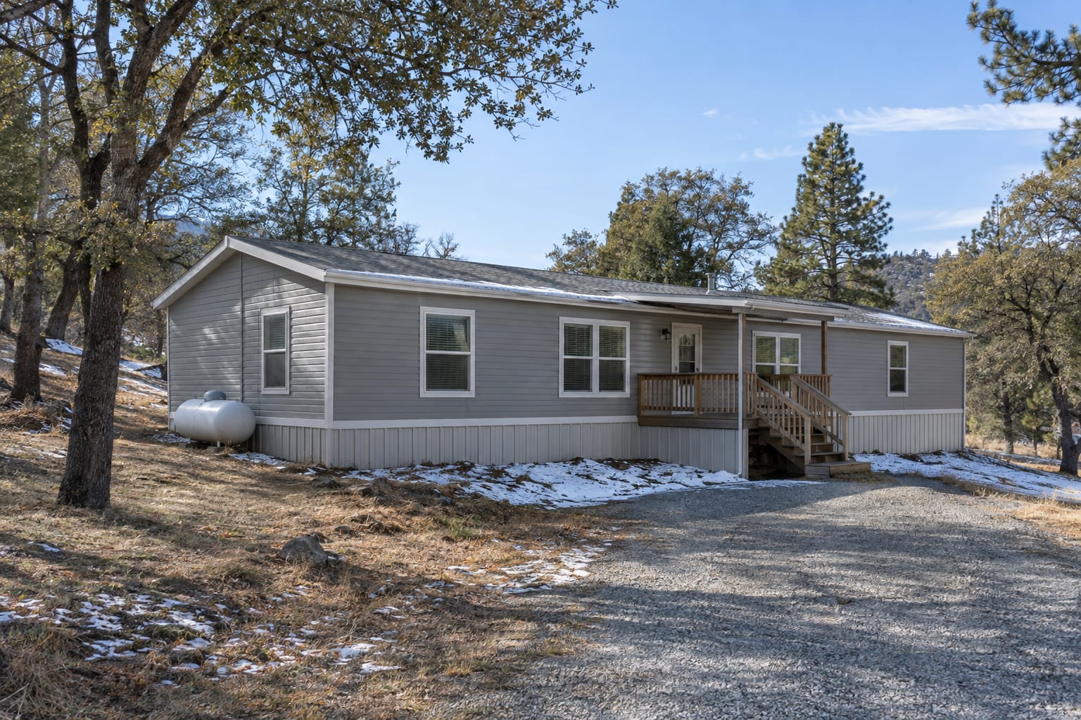 A double-wide manufactured home on a sloping mountain lot among oaks and pines, with patches of snow on the ground and a propane tank beside it.