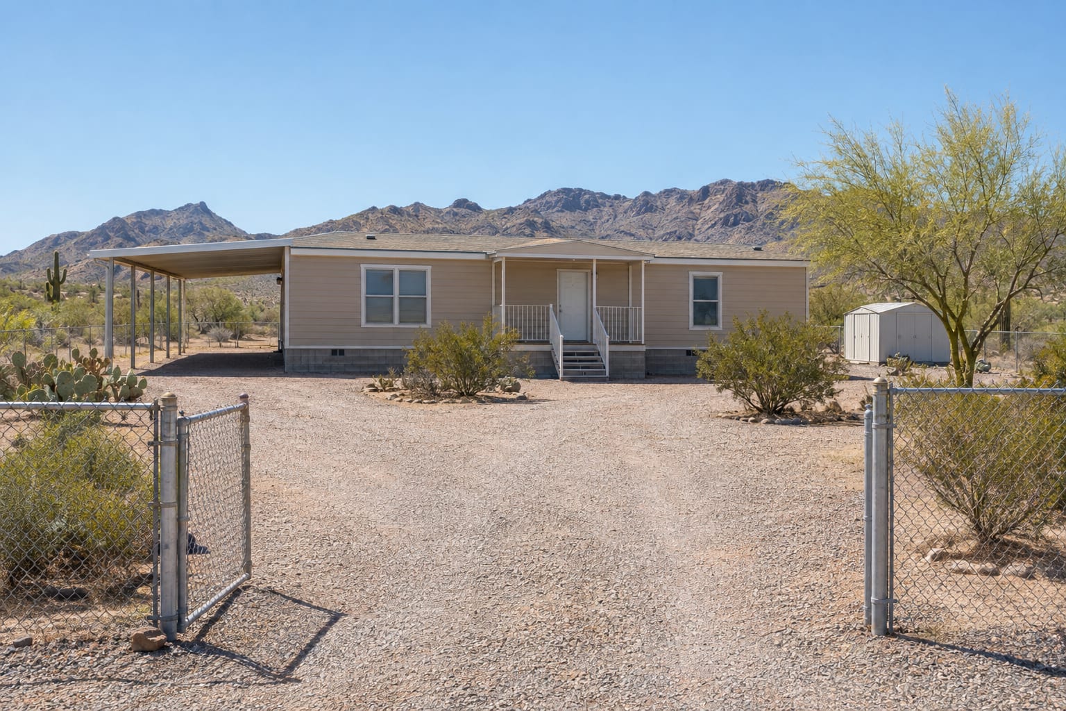 A manufactured home with a metal carport on its own fenced lot in the Arizona desert, seen from the end of a long gravel driveway with mountains behind