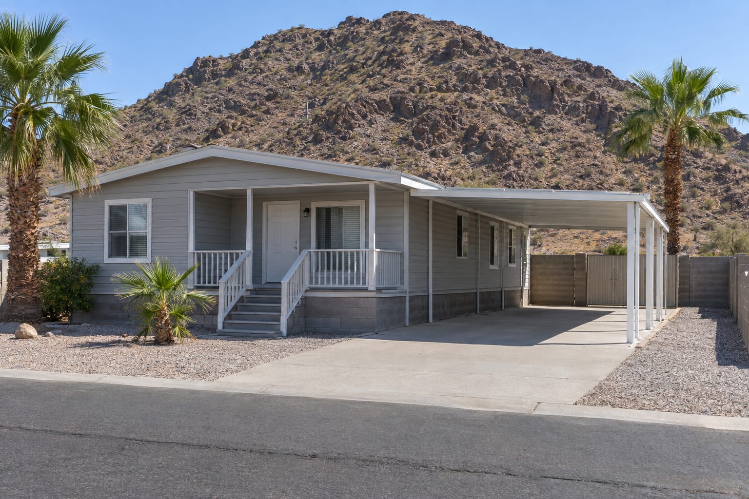 A double-wide manufactured home on its own walled lot with a carport and a rocky hillside behind it in a desert river town.