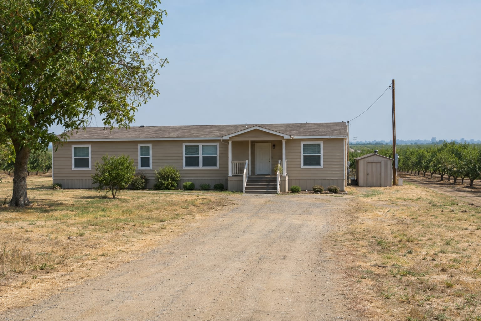 A double-wide manufactured home on a rural parcel beside rows of orchard trees, with a small pump house and a power pole.