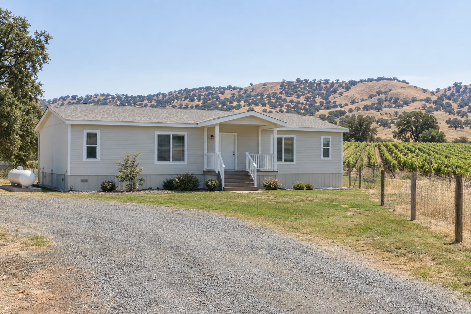A manufactured home with skirting on a flat rural parcel, with a propane tank beside it, a gravel driveway and rows of grapevines in the distance