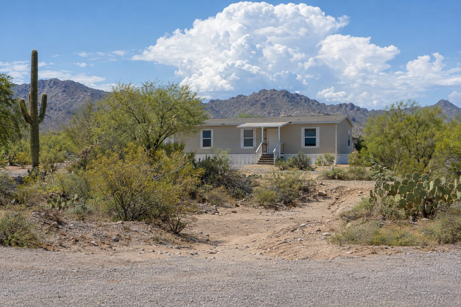 A dry sandy wash running down to a gravel road in the Tucson area, with a manufactured home beyond the desert brush and monsoon clouds building over the mountains.