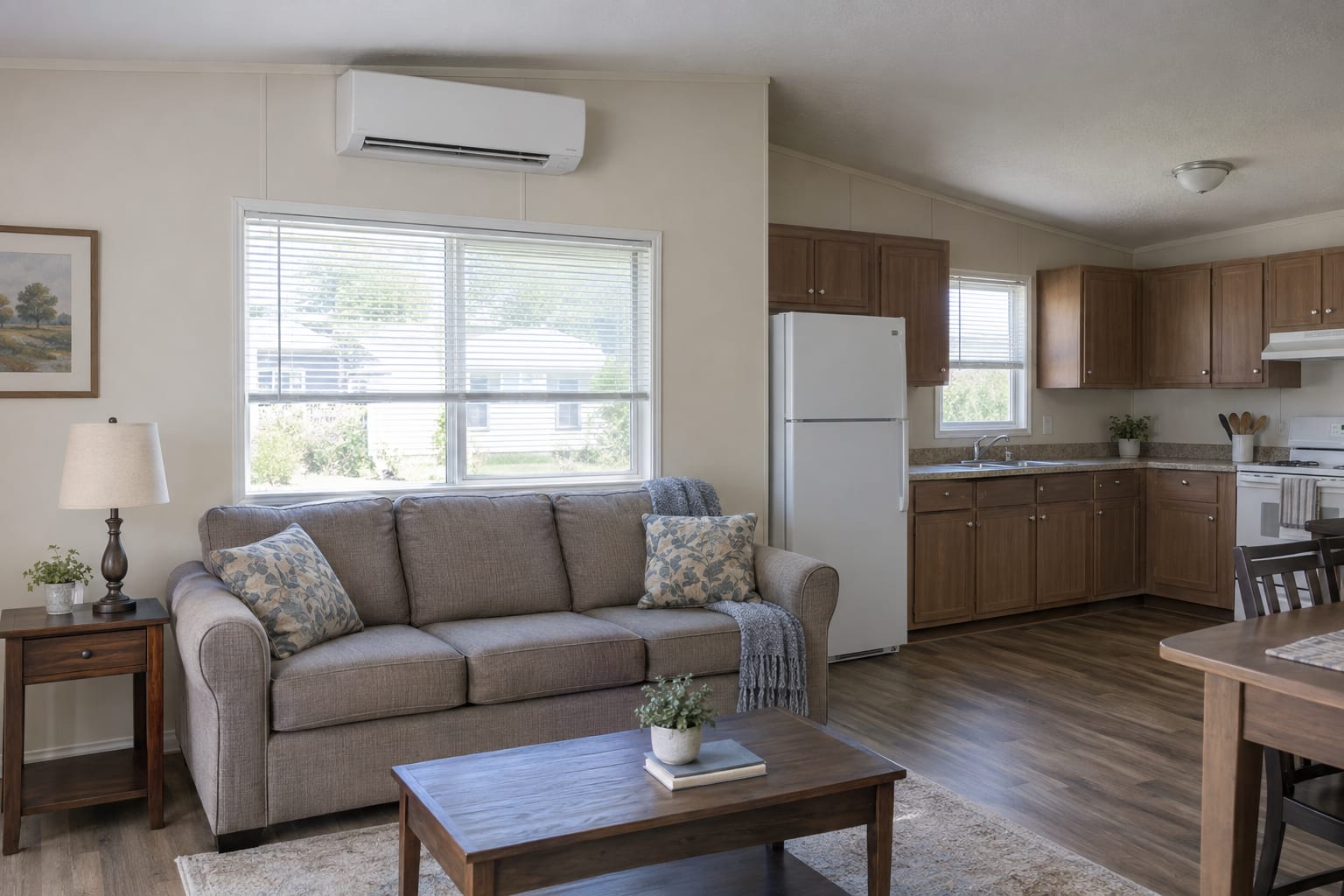 A ductless mini-split indoor unit mounted high on the wall of a manufactured home living room above a window, with a sofa below.
