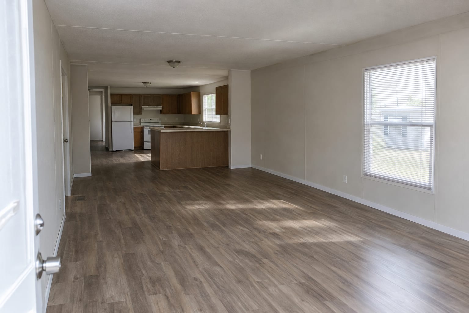 A clean, empty living room in a manufactured home with wood-look floors and a window with white blinds, seen from the entry