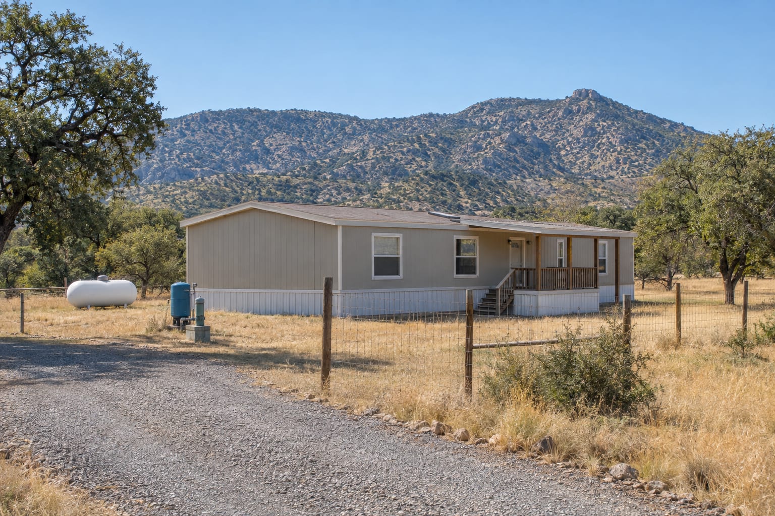 A manufactured home on a fenced high-desert grassland parcel in southeastern Arizona, with a propane tank, a well head and a gravel drive, oak-dotted mountains behind.