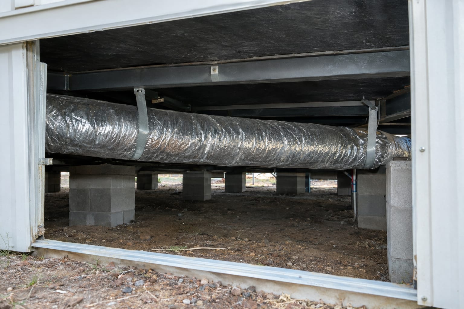 A large insulated flexible duct strapped up beneath the steel frame of a double-wide manufactured home, seen through an opening in the skirting, with the ground below it dry.
