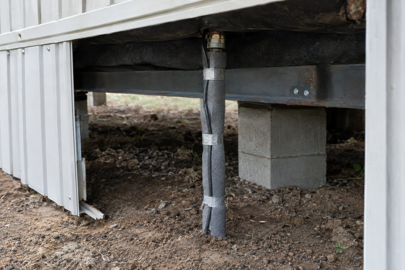 An insulated water supply pipe wrapped with heat cable rising from the ground beneath a manufactured home, seen through an open skirting panel.