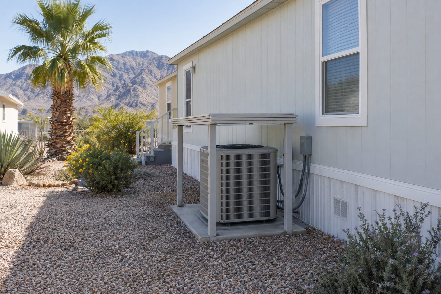 A ground-mounted air-conditioning condenser under a small shade cover beside the skirting of a manufactured home in a desert community, gravel yard and fan palm nearby