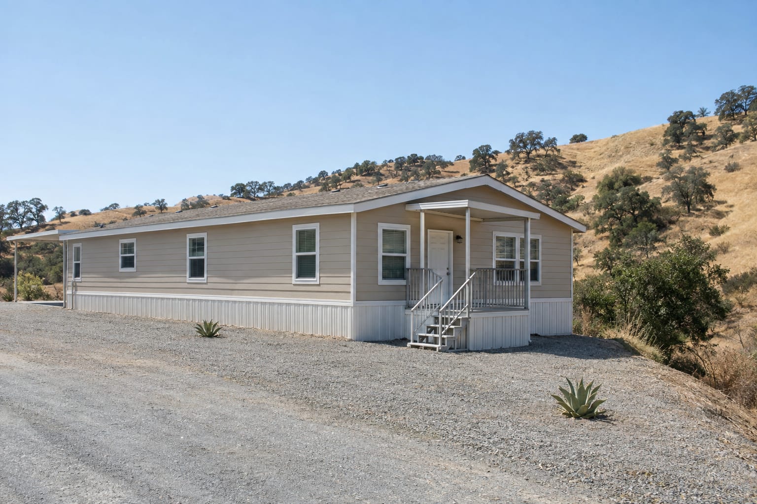 A manufactured home at the edge of dry chaparral hills, with a wide band of cleared gravel between the home and the brush