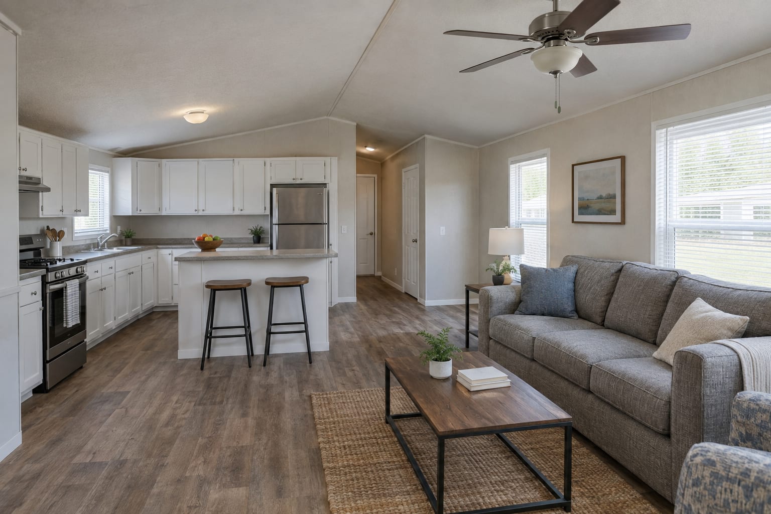 The open kitchen and living area of a double-wide manufactured home, with white cabinets, a small peninsula and wood-look plank flooring