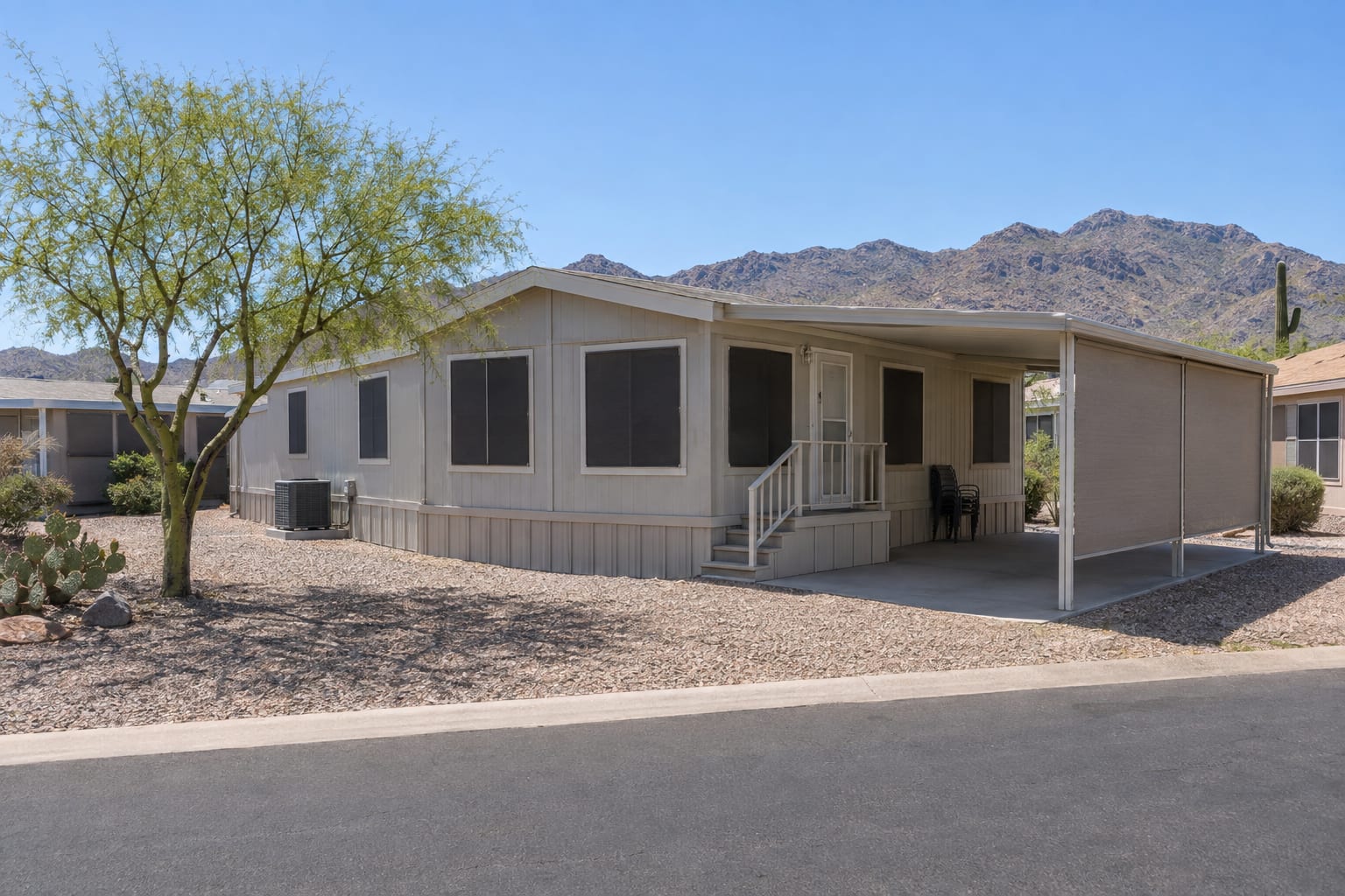 A double-wide manufactured home in midsummer with dark solar screens on the windows, a shaded empty carport and an air-conditioning unit beside it.