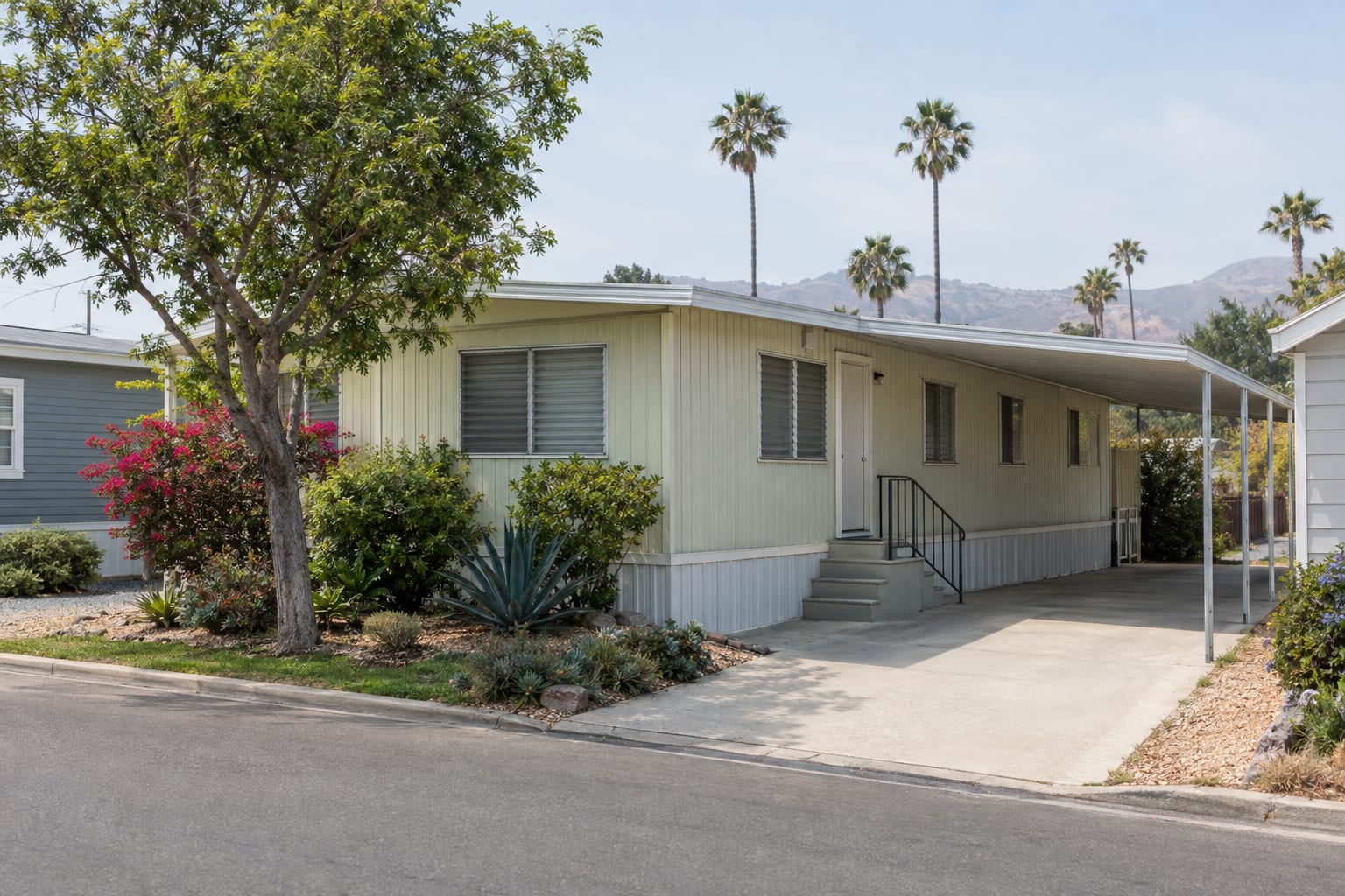 An older manufactured home with aluminum siding, a long metal carport awning and mature shrubs in an established community
