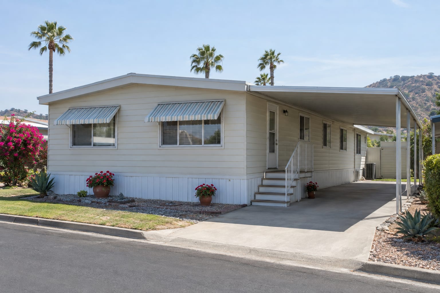 A well-kept older manufactured home with pale aluminum siding, window awnings and a long carport on a community street