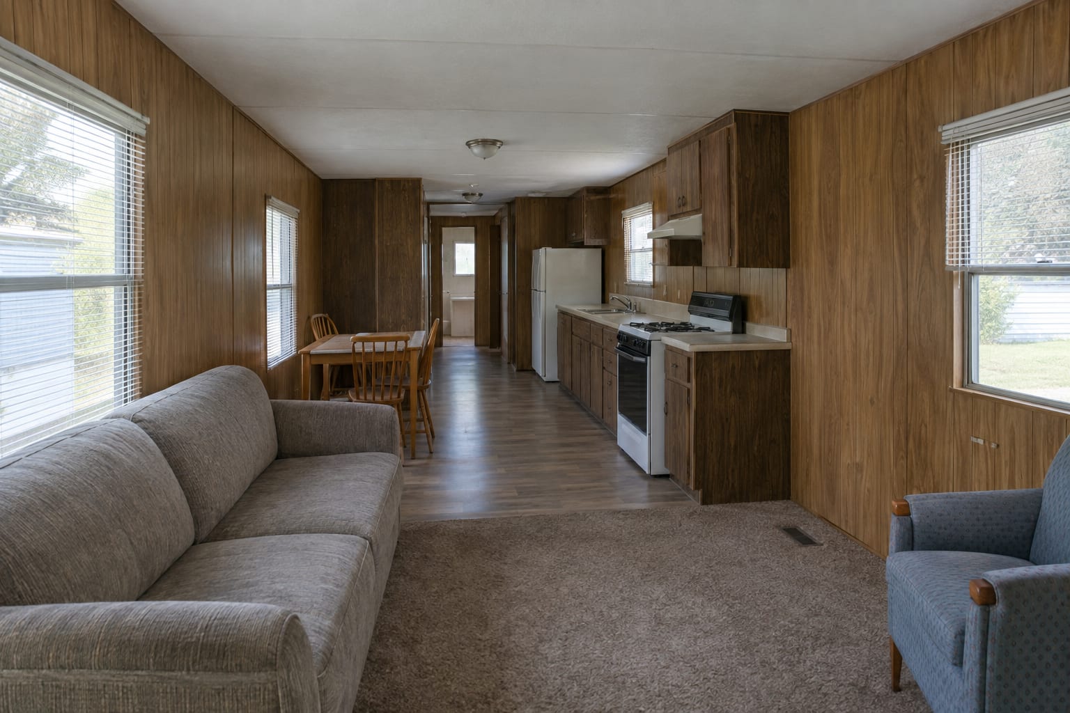 The narrow living room and galley kitchen of an older single-wide manufactured home, with wood-grain wall paneling and windows on both sides.