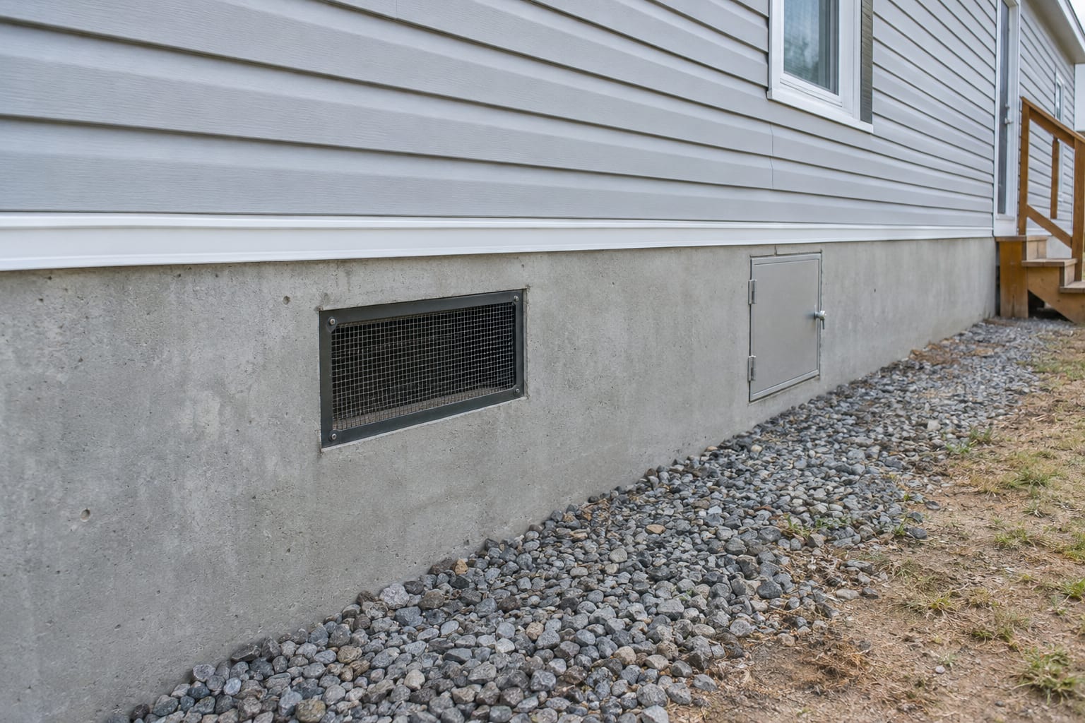 A low view along the base of a manufactured home on a permanent perimeter foundation, showing a concrete stem wall with a screened crawl-space vent below the siding.