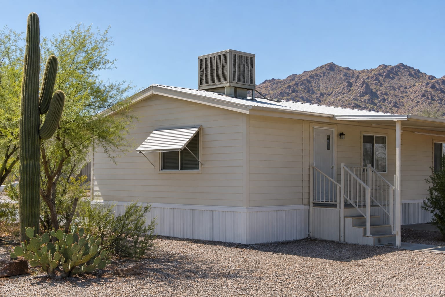 An evaporative cooler mounted on the white-coated roof of a manufactured home, with a saguaro and desert mountains behind.