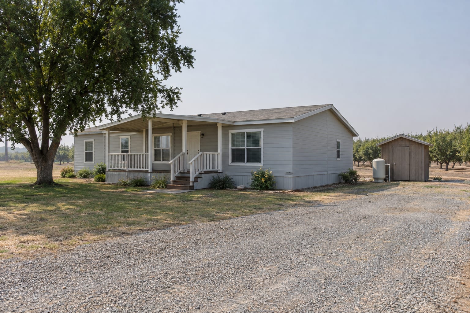A double-wide manufactured home on a rural valley parcel with a small well pump house in the yard and orchard rows behind.