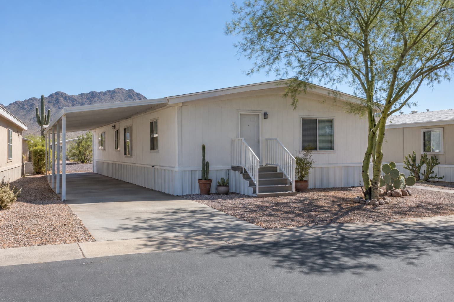 A single-wide manufactured home with an awning carport on a gravel space in an Arizona community, with neighboring homes close by.