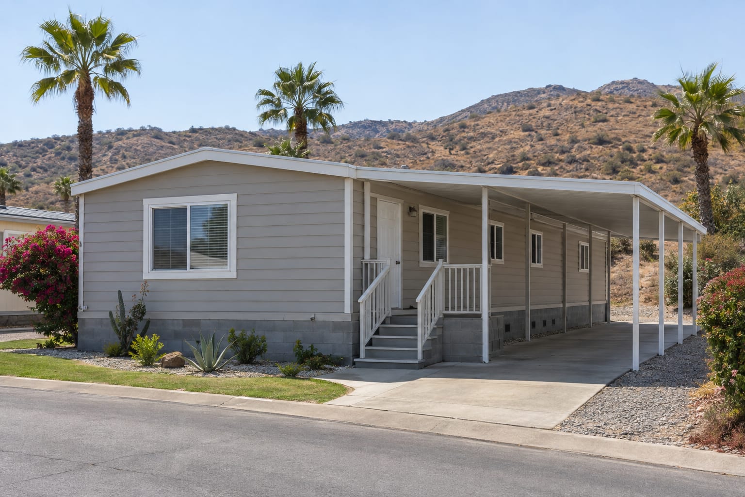 A single-wide manufactured home with concrete-block skirting and a metal carport at the edge of a foothill community, with a dry chaparral slope rising behind