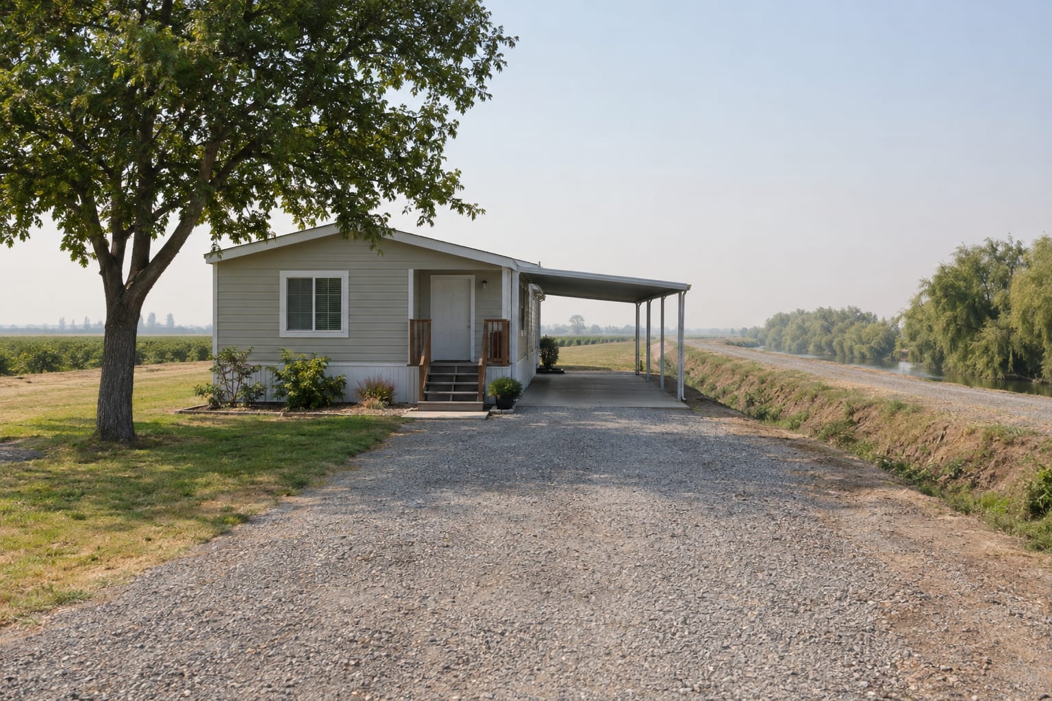 A single-wide manufactured home with a carport on a flat lot beside a levee road lined with willows, with a slough beyond.