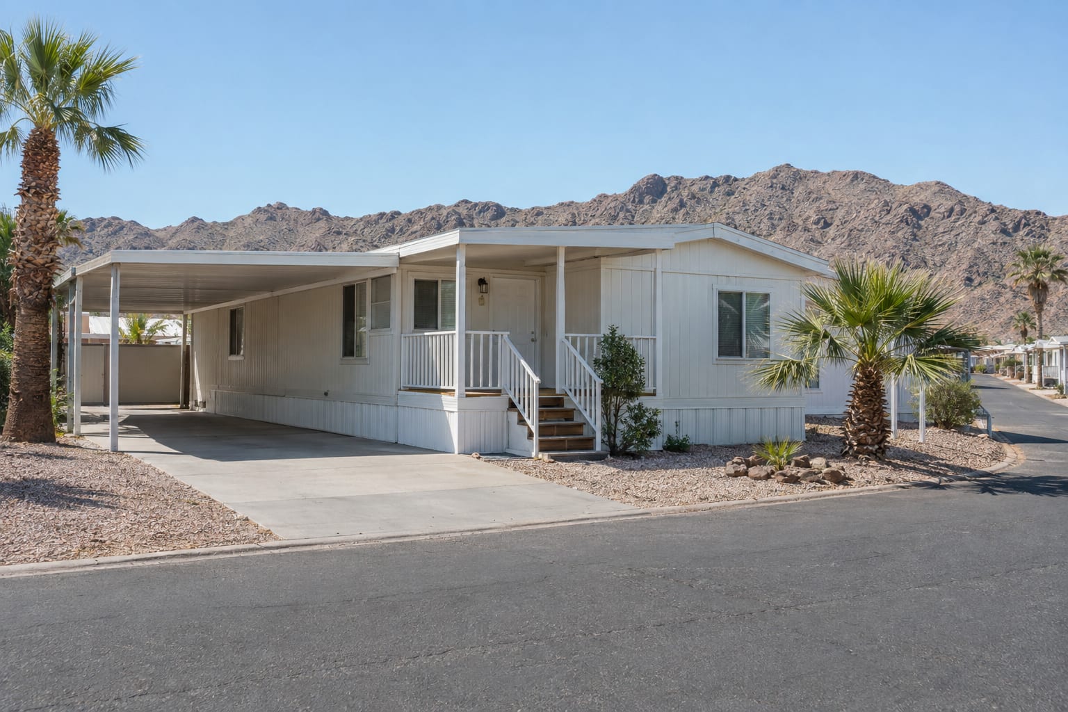 A manufactured home with a carport and a small porch on a community street in a desert river valley, with bare mountains behind.