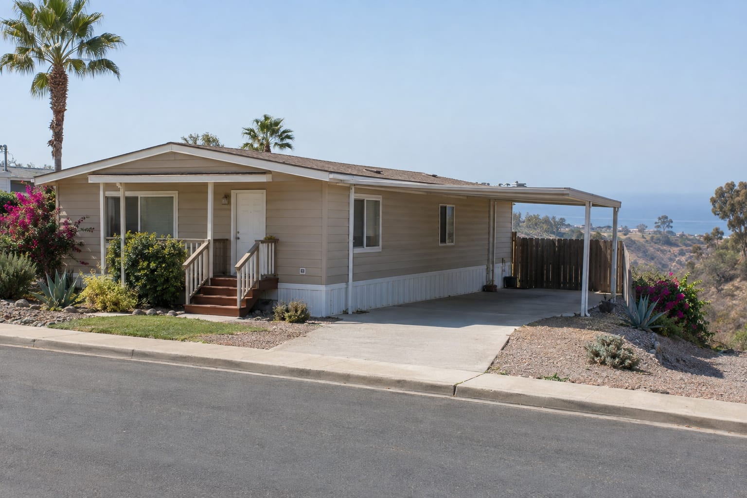 A manufactured home with a carport on a flat community street, with a brush-filled canyon dropping away behind the lot