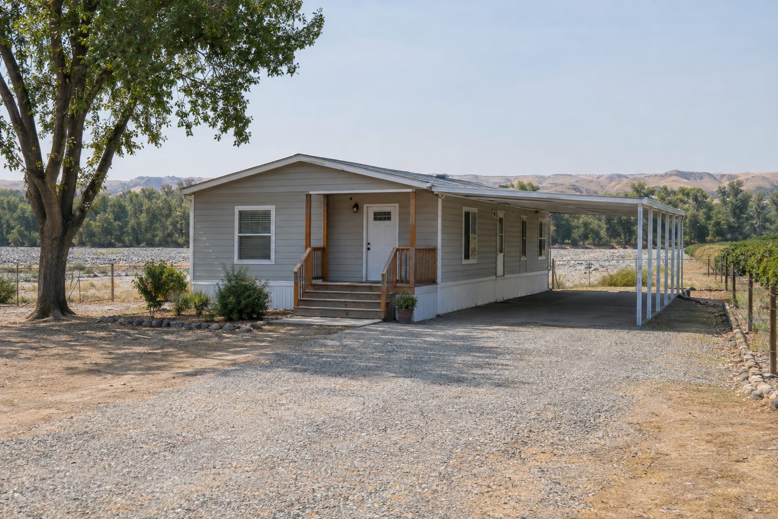 A single-wide manufactured home with a carport on a flat lot, with the dry gravel bed of a valley river and a line of cottonwoods behind it.