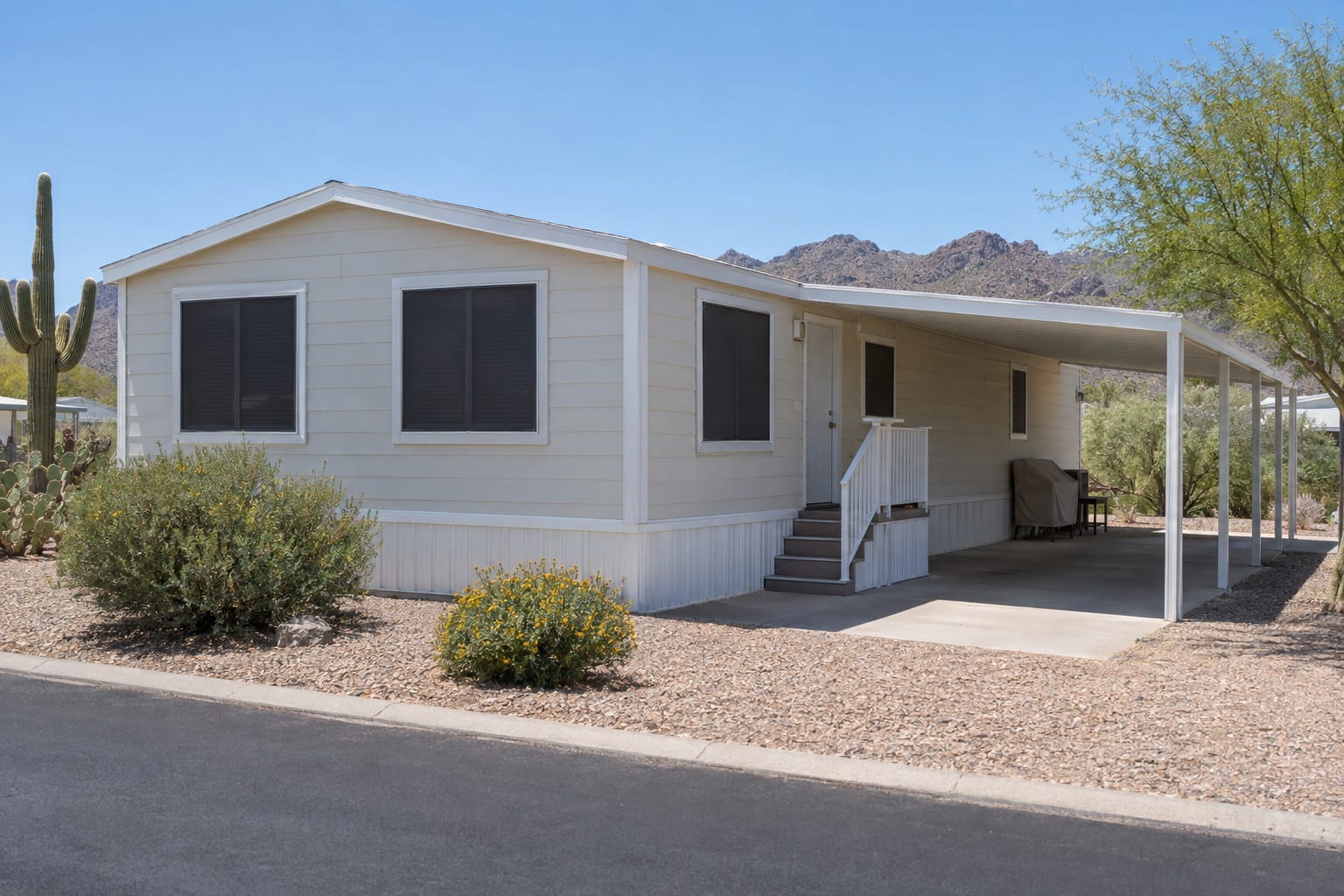 A manufactured home with dark sun screens on the windows and an empty carport, with covered patio furniture stacked against the wall under it.