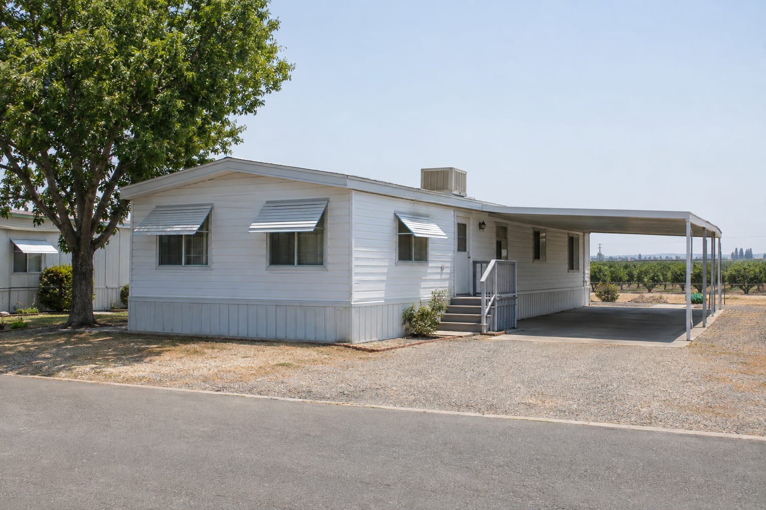A single-wide manufactured home with a rooftop evaporative cooler, window awnings and a shaded carport on a community street in summer light.