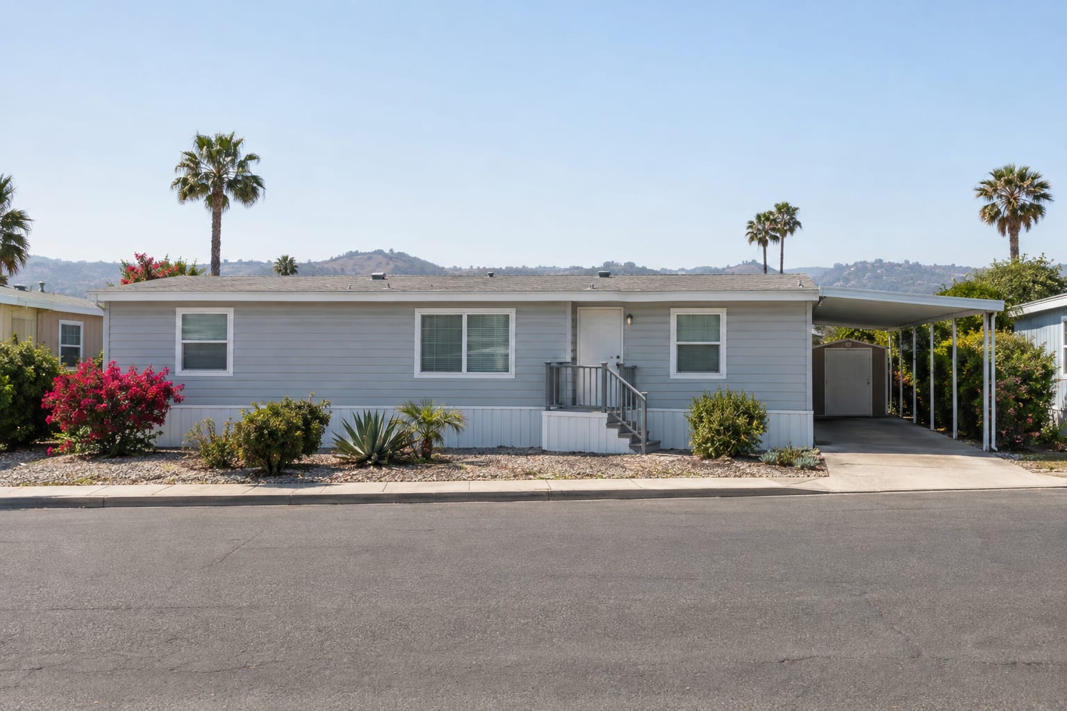 A manufactured home seen along its full length from across a community street, with entry steps, skirting and a carport.