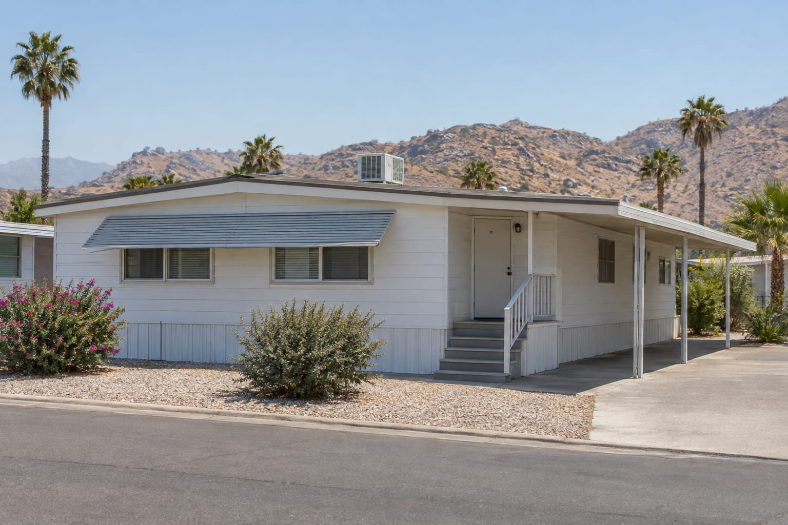 A single-wide manufactured home with a rooftop air-conditioning unit and a shade awning over its windows on an inland valley community street, dry hills behind
